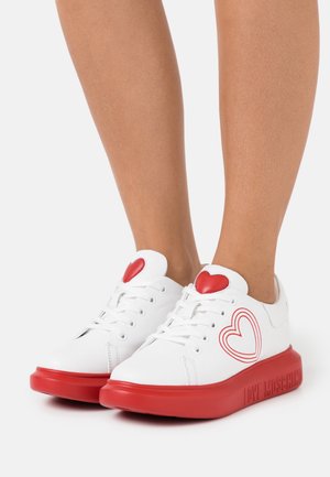 Witte sneakers met rode zolen, een rood hart op de tongen en een rood omlijnd hart aan de zijkanten, gedragen op blote benen. "LOVE MOSCHINO" gegraveerd op de zool.