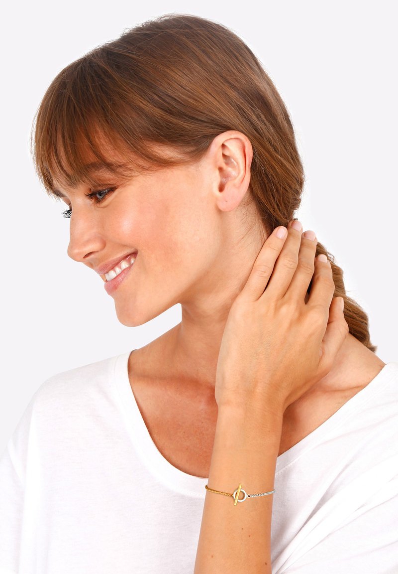 Arany és ezüst fonott karkötő, kapoccsal, amely egy csuklón van viselve, egyszerű kör alakú dizájnnal. A modellen fehér ing van.