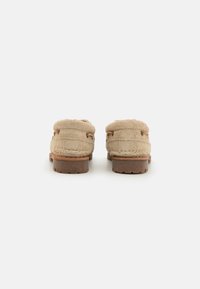 Zapatos slip-on de ante beige con forro suave, forma redondeada, suela de goma y acentos en tono marrón claro. Presentan costuras dobles y un detalle de hardware mínimo.