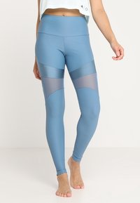 Modré legíny s vysokým pasem, které mají hladký povrch, síťované panely po stranách a lesklé horizontální akcenty. Vyrobeny z pružné látky.