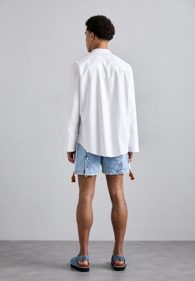 Chemise blanche à manches longues avec une coupe droite, associée à un short en denim bleu clair avec des boutons décoratifs et des pompons, et des sandales décontractées bleues.
