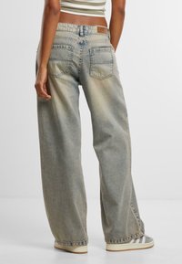 Light blue wide-leg jeans feature a faded wash, two back pockets, and a brown leather patch on the waistband. Worn with gray sneakers.