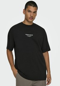 Svart bomulls t-shirt med avslappnad passform, rund halsringning och korta ärmar. Har "KURATERADE GRUNDER" tryckt i vitt på bröstet.