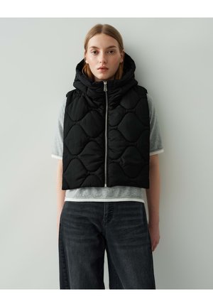 Zwarte gewatteerde korte puffer vest met capuchon, voorzien van een rits aan de voorkant en een golfpatroon, gelaagd over een grijs t-shirt met korte mouwen.