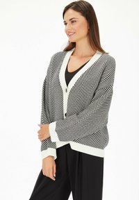 Czarny i biały kardigan w wzór jodełki z dekoltem w kształcie V, zapięciem na guziki i ściągaczami przy rękawach. Miękki dzianinowy materiał, luźny krój i lekka konstrukcja.