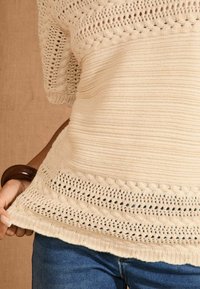 Maglione lavorato a maglia di colore crema con motivi testurizzati, bordi in pizzo e vestibilità morbida. Tenuto a mano all'orlo sopra jeans blu scuro.