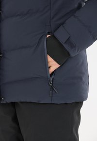 Marineblaue Pufferjacke mit glänzender Oberfläche, ausgestattet mit seitlichen Reißverschlusstaschen und elastischen Bündchen für einen engen Sitz. Strukturiertes Gewebe und Isolationsdetails sind sichtbar.