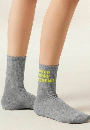 Pieds portant des chaussettes grises côtelées, l'une d'elles affichant en jaune sur la cheville le texte "IL ME FAUT PLUS DE WEEK-END".