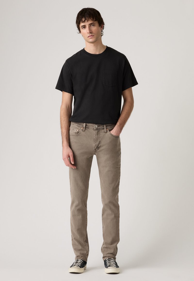 Giovane uomo in piedi, indossa una maglietta nera, jeans beige e scarpe da ginnastica bianche e nere, su sfondo chiaro e uniforme.