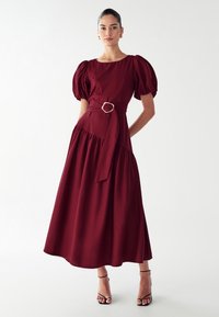 Bordeaux midi-jurk met pofmouwen, aansluitend lijfje en een ceintuur in de taille met een ronde metalen gesp. Lichtgewicht stof, zachte textuur.