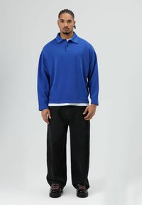 Blauwe polo sweatshirt met een kraag en twee knopen, gecombineerd met zwarte wijde broeken en zwarte schoenen met roze veters. Effen achtergrond.