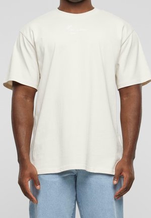 Homme portant un t-shirt ample à manches courtes de couleur blanc cassé uni, associé à un jean bleu clair, debout devant un fond uni.