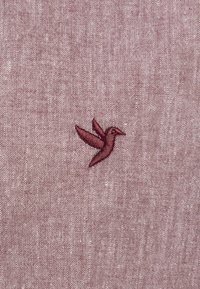 Colibri brodé en fil bordeaux foncé sur un tissu texturé, doux et rose clair. Le design simple met l'accent sur la forme de l'oiseau et ses ailes.