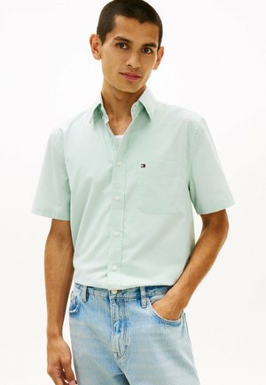 Junger Mann mit kurzen Haaren, der ein mintgrünes, kurzärmeliges Hemd mit Knöpfen und hellblaue Jeans trägt, steht mit einer Hand in der Tasche.