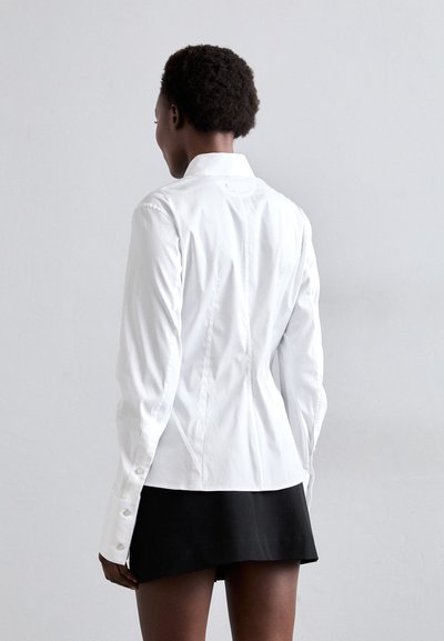 White button-up shirt with long sleeves, featuring a fitted design, collared neckline, and subtle back seam details, paired with a black skirt.