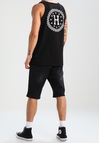 Top de tirantes negro de algodón con un logotipo circular blanco que presenta una "H" y estrellas en la parte posterior, combinado con pantalones cortos de mezclilla negros y zapatillas negras.