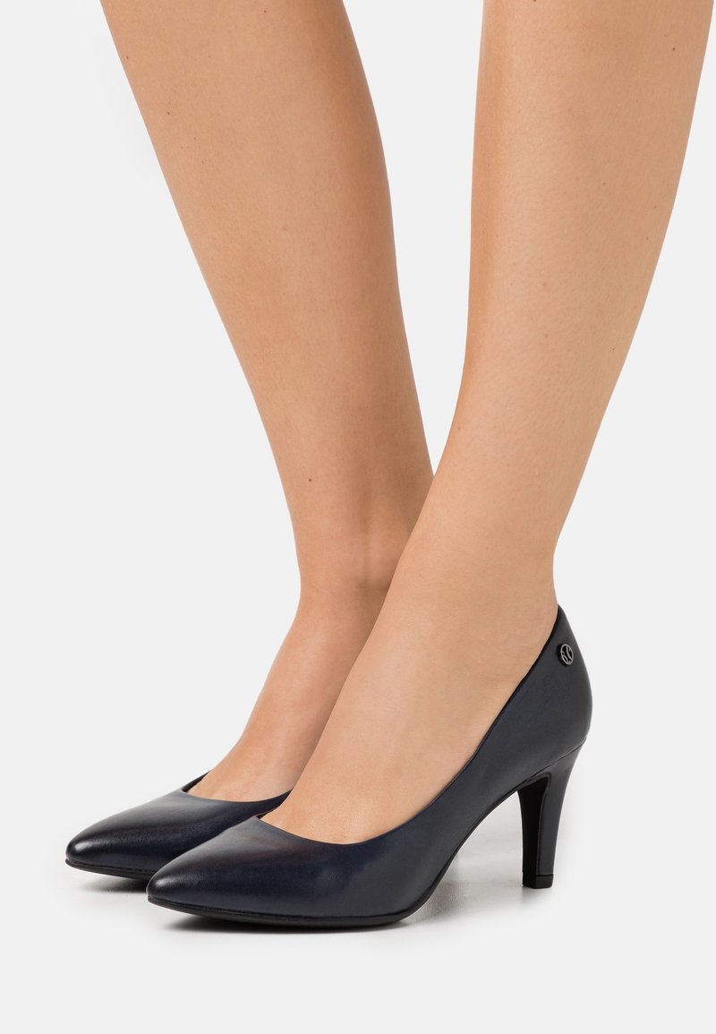 Femme portant des escarpins noirs en cuir à bout pointu avec un talon de hauteur moyenne, debout sur une surface blanche unie.