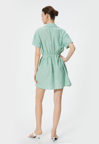 Grøn- og hvidstribet skjortekjole med korte ærmer, indsnævret talje, åben krave og flagrende nederdel. Bæres med minimalistiske sandaler.