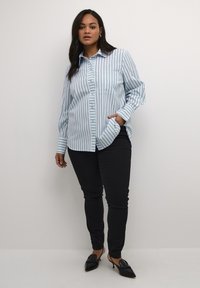 Camisa de botones azul claro con rayas verticales. Tiene un cuello con solapa, un bolsillo en la parte frontal y mangas largas. Se lleva con pantalones ajustados negros.