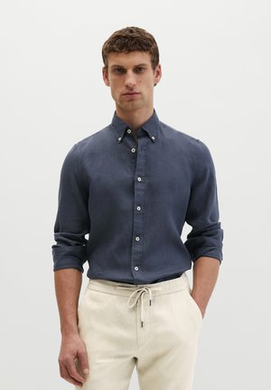 Mann trägt ein dunkelblaues Hemd mit Knöpfen und aufgekrempelten Ärmeln sowie beige Hose mit Kordelzug, steht vor einem schlichten hellen Hintergrund.