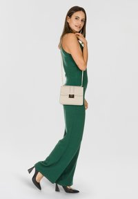 Borsa beige con tracolla a catena, forma rettangolare, superficie testurizzata e chiusura a flap con fermaglio dorato. Indossata con una tuta verde.