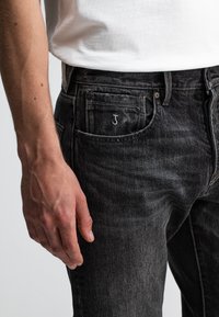Zwarte denim jeans met een slim fit, voorzien van een zichtbaar geborduurd 'J' op de zak en lichte slijtage. Hand rustend bij de tailleband.