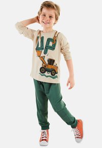 Beige Langarmshirt mit grünen Grafikbuchstaben und einem orangen Baggerdesign. Dunkelgrüne Hose mit elastischem Bund und orangefarbene Sneakers.