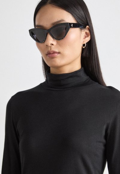 Occhiali da sole neri a forma di occhio di gatto con lenti scure, montatura nera testurizzata e accenti di glitter discreti. Il modello indossa un dolcevita nero.