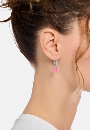 Rechtes Ohr mit silbernem Creolenohrring, der kleine Diamanten und einen hängenden pinkfarbenen Gummibärchen-Anhänger zeigt, vor hellem Hintergrund.