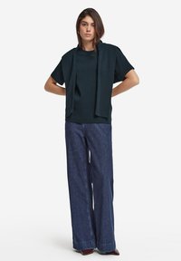 Mörkgrön kortärmad skjorta med en matchande slips vid halsen, i kombination med vida denimbyxor med en fåll.