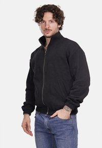 Giacca nera con zip realizzata in un morbido tessuto testurizzato. Presenta un colletto alto e polsini elastici, con tasche laterali e una vestibilità rilassata.