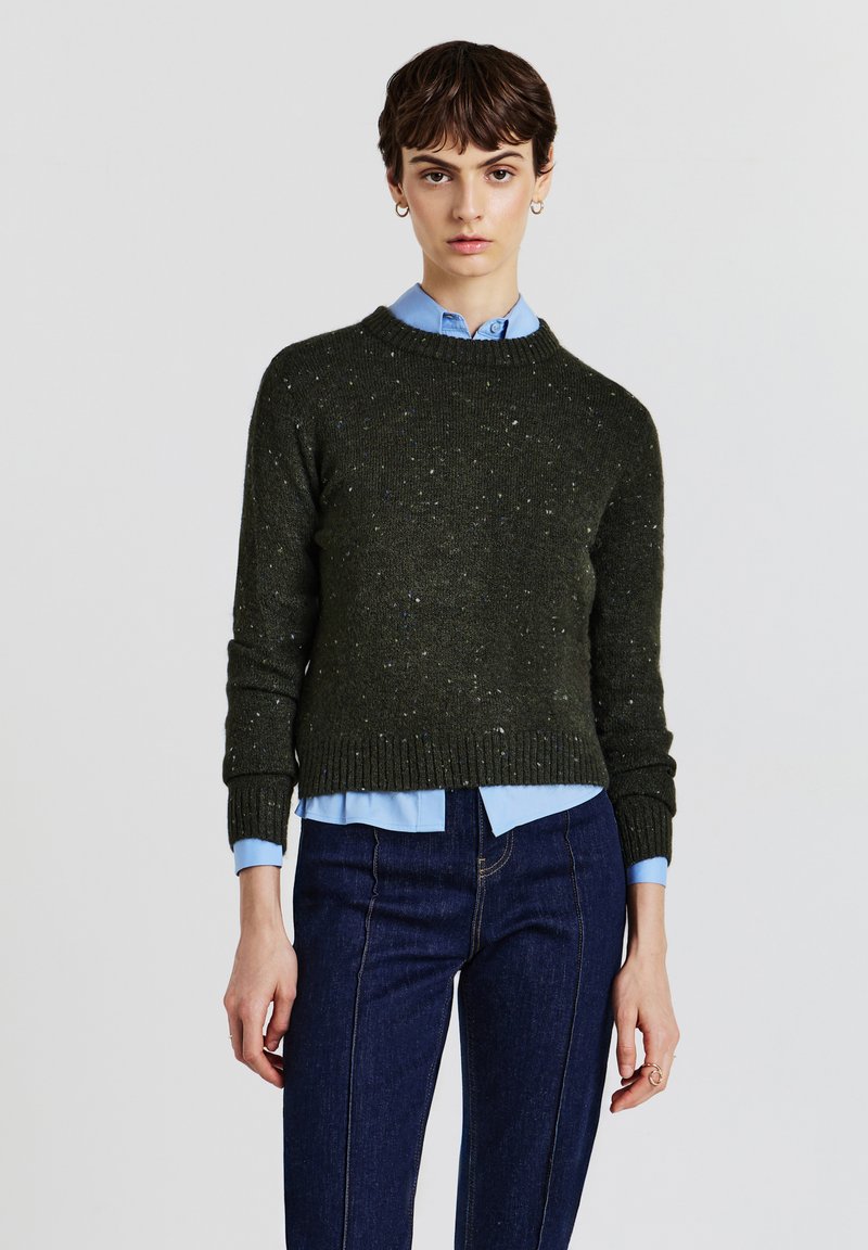 Grün gesprenkelter Strickpullover mit Rundhalsausschnitt, gerippten Bündchen und Saum, über einem hellblauen Hemd mit Kragen gelegt, kombiniert mit dunklen Jeans.