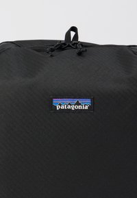 Juodas kuprinė, pasižyminti tekstūruota paviršiaus, raišteliu uždaru ir Patagonia logotipo patch'u su kalnų dizainu mėlynais ir purpuriniais akcentais.