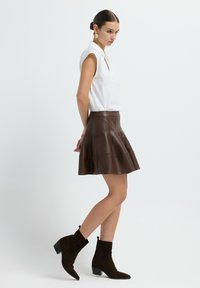 Jupe évasée en faux cuir marron avec des détails de couture, associée à un blouse blanche à manches courtes et des bottines marron foncé.