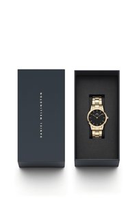 Reloj de pulsera dorado con esfera negra y acentos dorados, presentado en una caja azul oscura con un interior suave y negro. Caja etiquetada "DANIEL WELLINGTON."