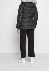 Chaqueta de plumas negra con capucha de pelo gris desmontable, diseño acolchado y mangas largas. Combinada con pantalones negros y zapatillas blancas.