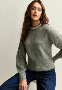 Grijze gebreide trui met een geribbelde zoom en wijde mouwen, met een bewerkte halslijn. Het materiaal lijkt zacht met een gladde textuur.