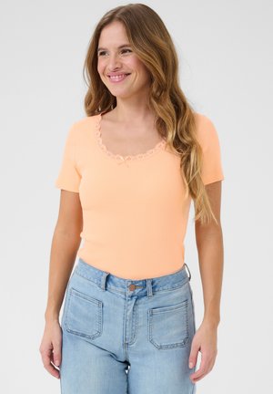 Pfirsichfarbenes Kurzarmoberteil mit einem gewellten Spitzen-Halsausschnitt und einer Schleifendekoration vorne, kombiniert mit hochgeschnittenen hellblauen Jeans.