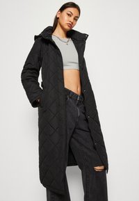 Cappotto lungo nero trapuntato con cappuccio, dotato di zip e chiusura a pressione, tasche laterali e una texture liscia. Indossato sopra una maglietta grigia e jeans scuri.