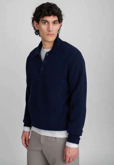 Giovane uomo con capelli ricci che indossa un dolcevita blu navy su una camicia bianca a maniche lunghe e pantaloni beige, in piedi davanti a uno sfondo semplice.