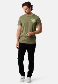 Olivenfarvet bomuld t-shirt med korte ærmer og rund halsudskæring, med et farverigt grafisk print på venstre bryst. Sorte jeans og hvide sneakers.