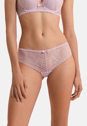 Vrouw met een lichtroze kanten beha en bijpassende kanten slip, zichtbaar van halverwege de romp tot de bovenbenen tegen een effen achtergrond.