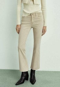Femme portant un pantalon évasé beige court avec ourlet brut, un pull côtelé crème et des bottines noires à talons, debout sur un sol vert clair.