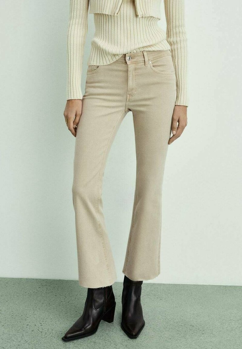 Femme portant un pantalon évasé beige court avec ourlet brut, un pull côtelé crème et des bottines noires à talons, debout sur un sol vert clair.