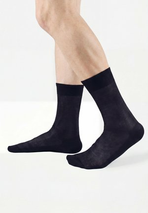 Beine mit halbtransparenten schwarzen Kniestrümpfen, die bis zur Mitte der Wade reichen, vor einem einfachen weißen Hintergrund stehend, ein Fuß leicht angehoben.