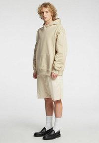 Beige hettegenser med frontlomme, kombinert med lyse beige shorts. Bruker svarte loafers og hvite sokker. Modellen står foran en ensfarget bakgrunn.