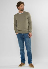 Jasnooliwkowy sweter dzianinowy z okrągłym dekoltem, w połączeniu z niebieskimi dżinsami i brązowymi butami. Prosty design i strukturalna tkanina.
