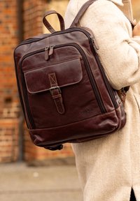 Brown leather backpack with adjustable straps, a front pocket, and a top handle. Features stitching accents and a sleek rectangular shape.