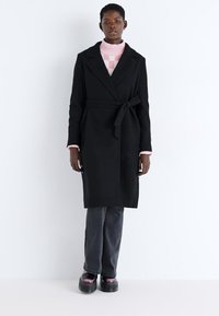 Black, knee-length wrap coat with a tie belt, tailored collar, and long sleeves over a pink checkered sweater and gray jeans.