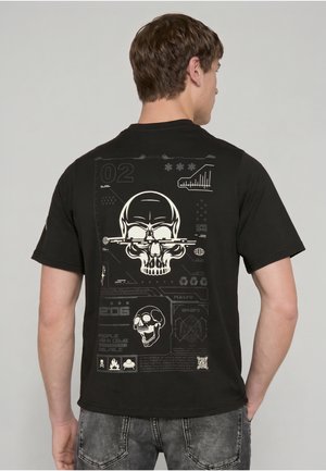 Mężczyzna w czarnej koszulce z białymi i szarymi futurystycznymi grafikami czaszki i cyfrowego interfejsu na plecach, stoi na jednolitym tle.